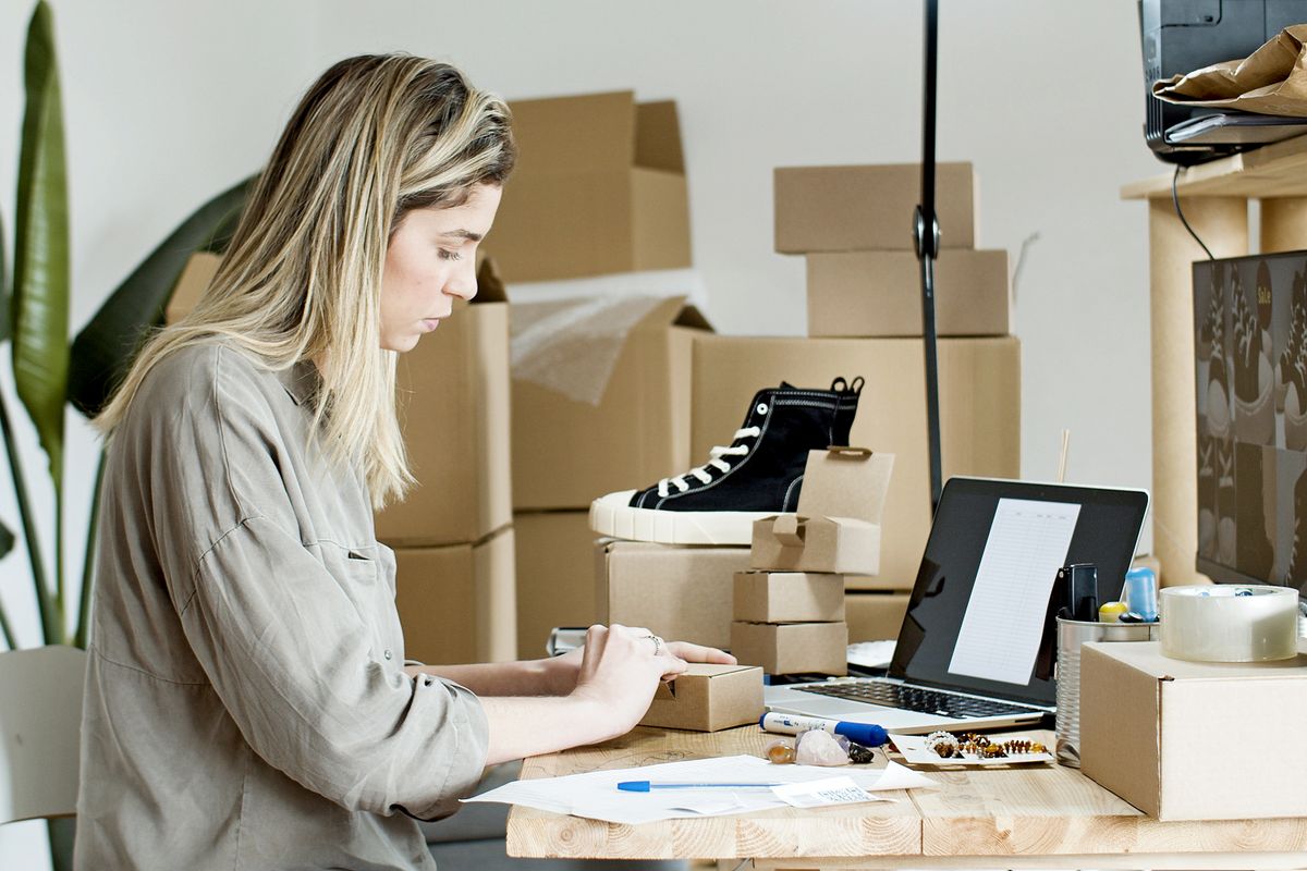 A person packing a parcel beside a laptop and stacked boxes.